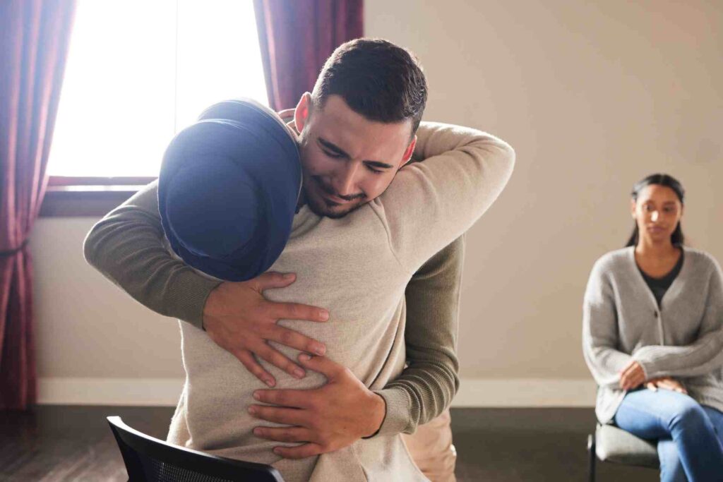 Man hugging another person during emotional mental health counseling session while woman sits in background
